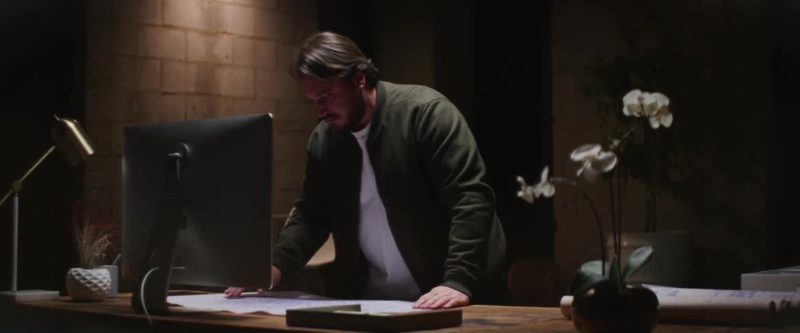 Man with beard green jacket and white shirt uses works on desktop in dim lit room