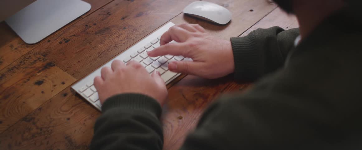A man types on a small wireless keyboard on a desk during the day