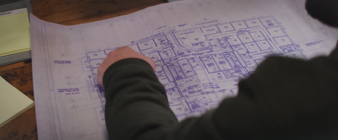 A man at a desk looking at blueprint plans tapping a section with his finger