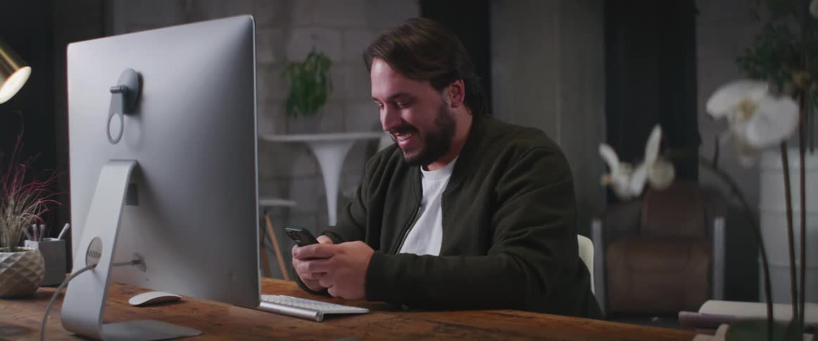 A man is sitting in front of a computer and smiling as he texts on his phone