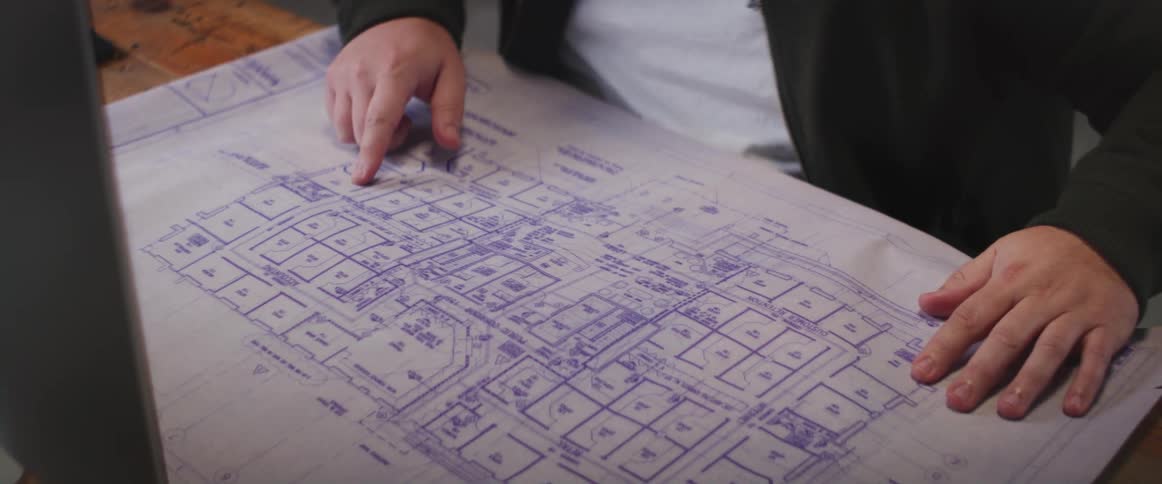 A man sitting at a computer desk retrieves a blueprint and unrolls it on the desktop