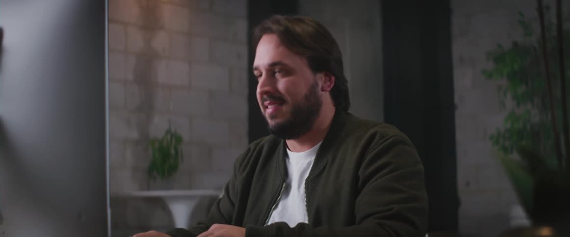 A man sitting at his desk puts his phone down and smiles at his computer monitor