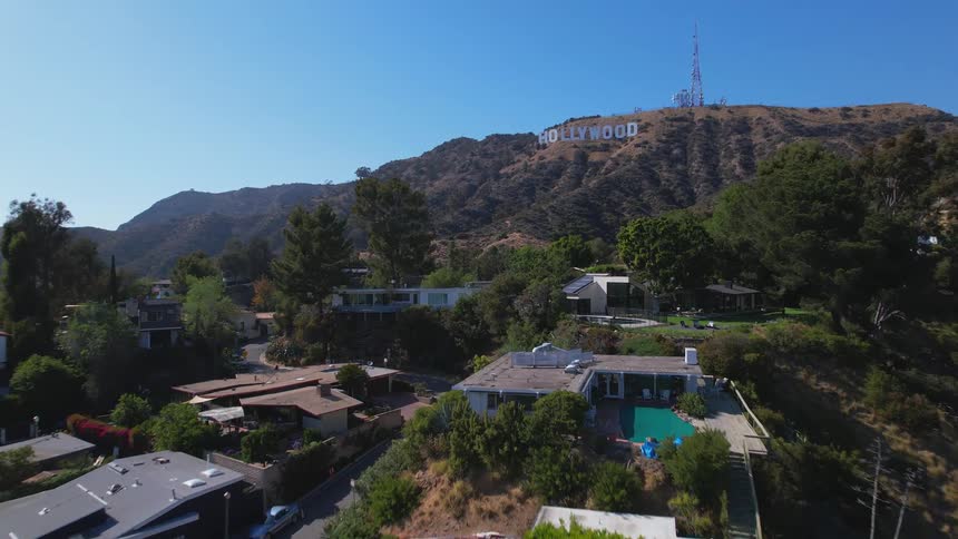 Aerial view of houses and trees in a hollywood neighborhood and the hollywood sign in the distance under a clear blue sky
