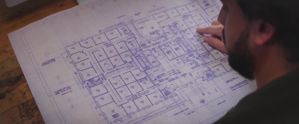 A man unrolls a blueprint atop a table and runs his finger along it
