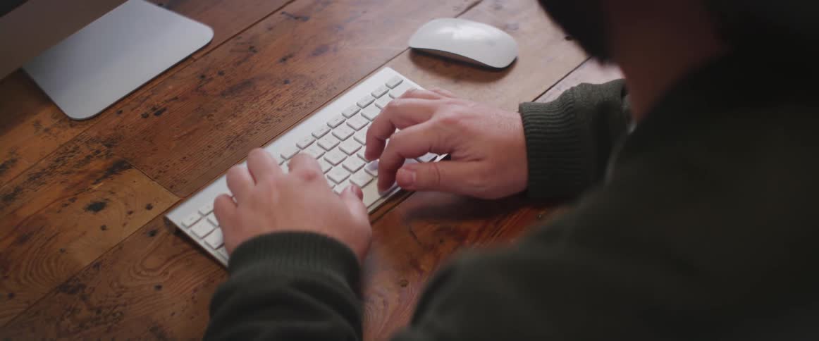Man wearing a long sleeved sweater types on a white keyboard as a white computer mouse sits to the right