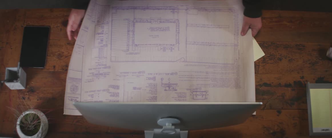 Someone unrolls a blueprint over a keyboard and mouse that are sitting on a desk and a hand is pointing to an area on the blueprint