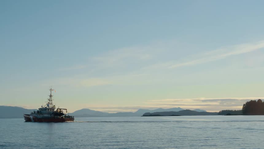 A coastguard ship with radar tower and antenna traveling past tree covered shore