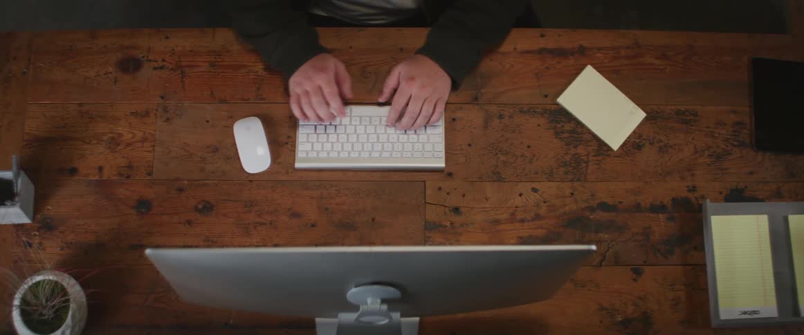 Person wearing long sleeve shirt sits at wooden desk touch typing on computer keyboard