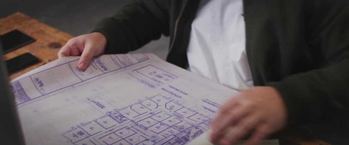 A man unrolls a blueprint on a computer desk while sitting indoors