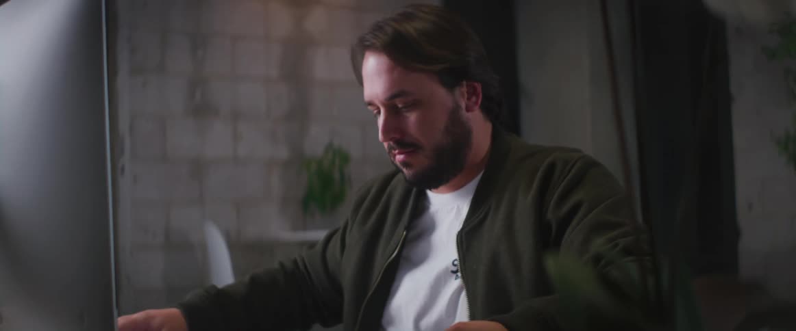 Man with beard green jacket and white shirt sitting at desk works on paper in front of computer screen