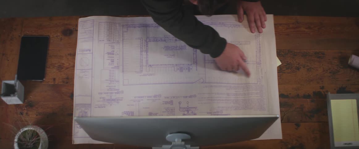A man is looking at a blueprint on the desk in front of a computer monitor and pointing to areas on the blueprint