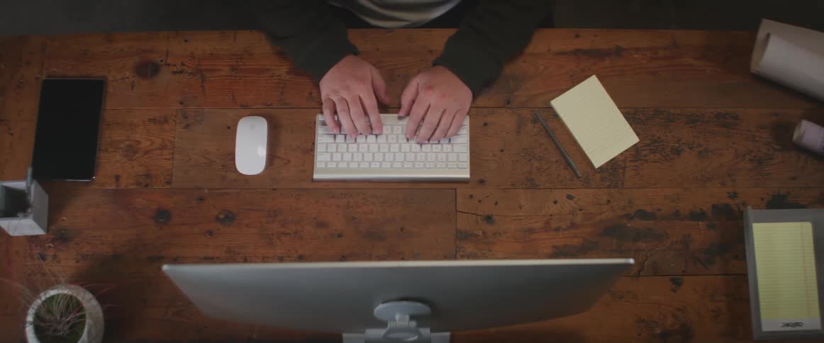 A person types on a wireless keyboard while sitting before a monitor at a wooden desk