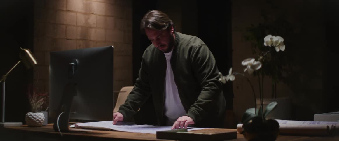 A man standing before a desk unrolls a blueprint on its surface