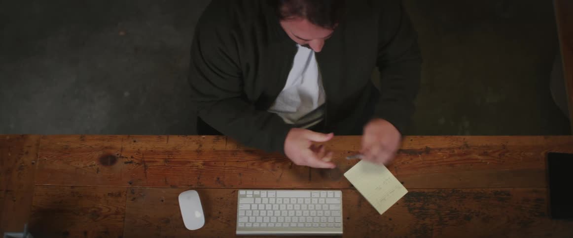 A man is sitting at a desk with a computer and he writes on paper and then starts typing on the keyboard