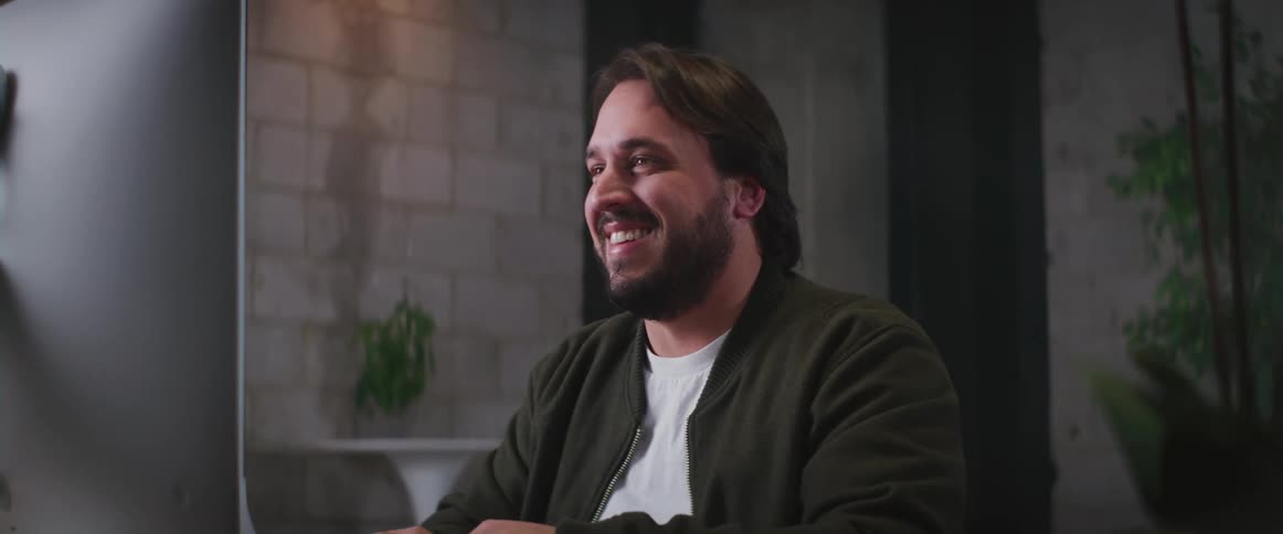 Man with beard green jacket and white t shirt sits at desk makes video call on computer smiling in room
