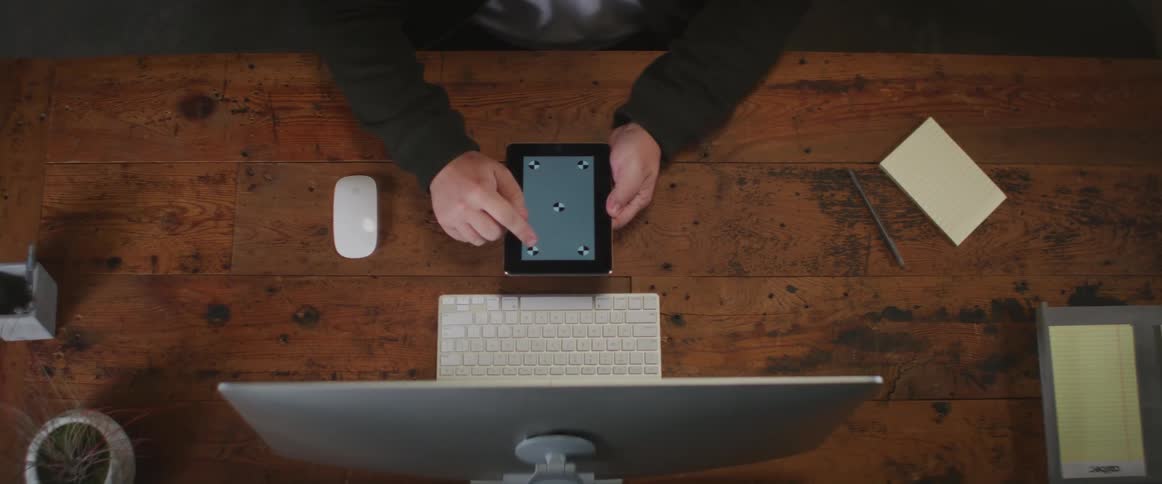 A man is sitting at a desk and he pushes the keyboard towards the monitor and uses a tablet computer