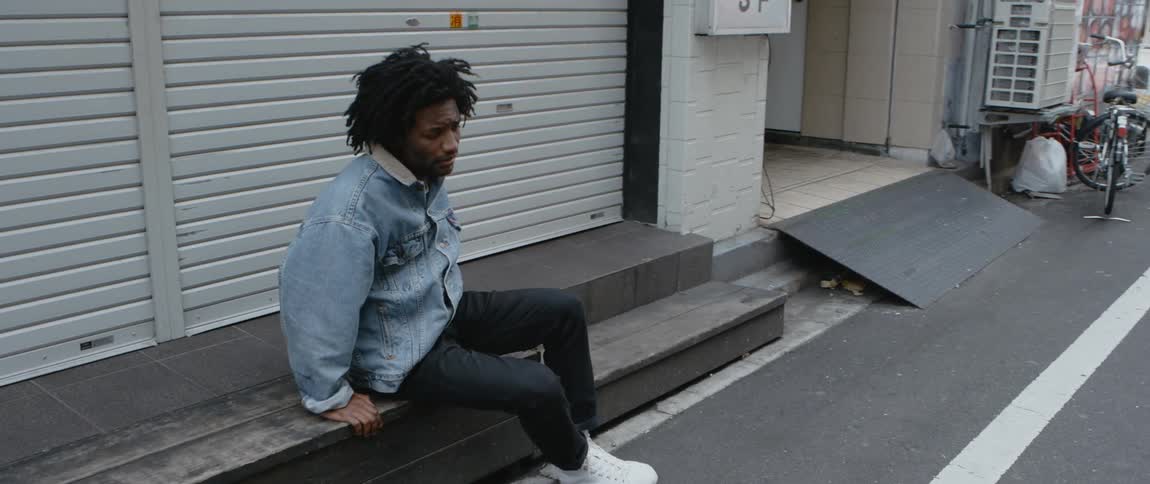 Man sits on a bench in front of a large door in an alley then pushes himself up to a standing position