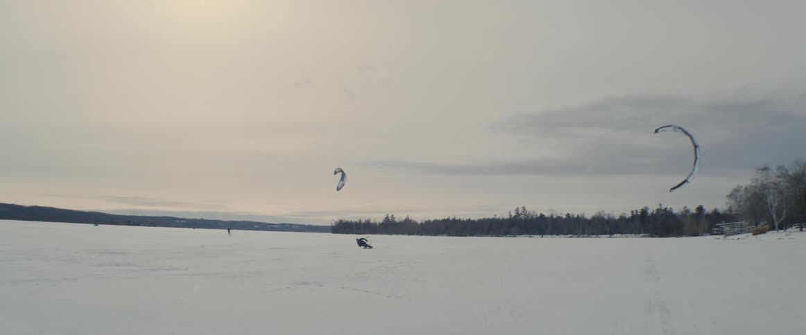 A kite skier leans far to the side of his skis as his kite speeds him over the snow covered ice