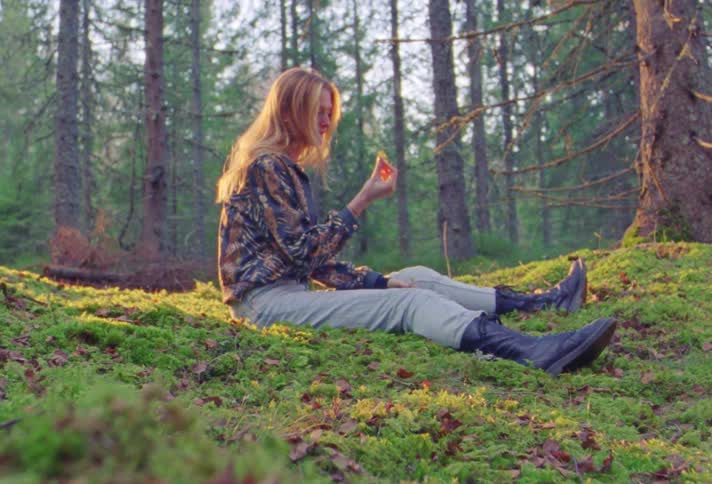 Middle aged blonde woman with blue blouse jeans and boots sits on ground and gets up in the woods