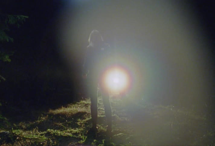 A woman stands in a dark field at night as a headlight starts coming towards her and she turns and shields her eyes from it as she looks