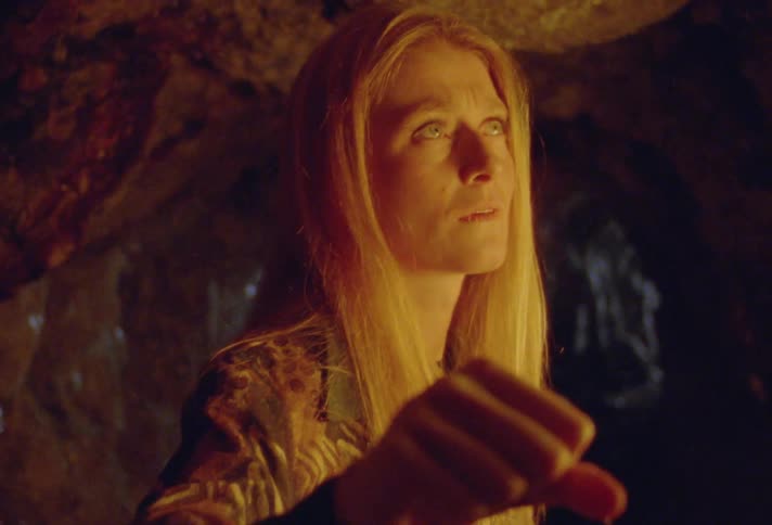 A woman with long blond hair is holding a lantern and looking around inside a cave and a light is shining at the end of the cavern