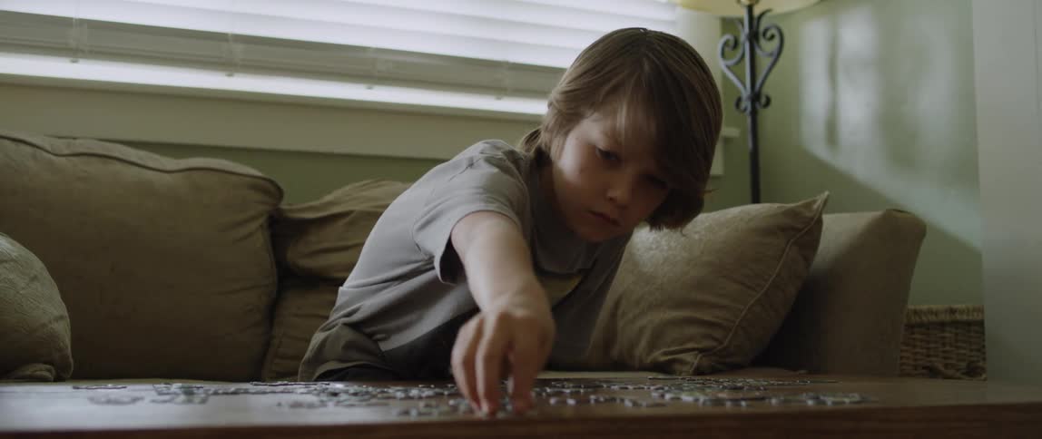 A young boy sits on a couch at home and picks up a piece of a puzzle and places it with another