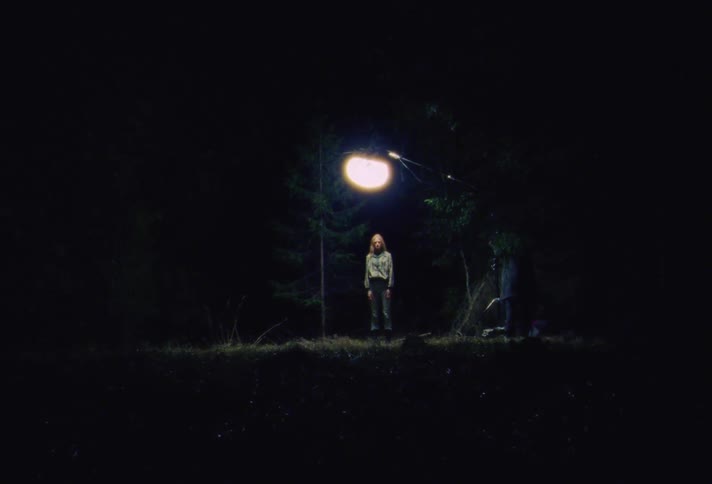Blonde woman stands under a bright light on a dark forest at night