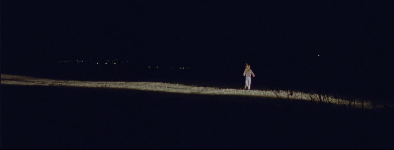 A woman is running out of woods at night and a light is shining on her as she runs across a field