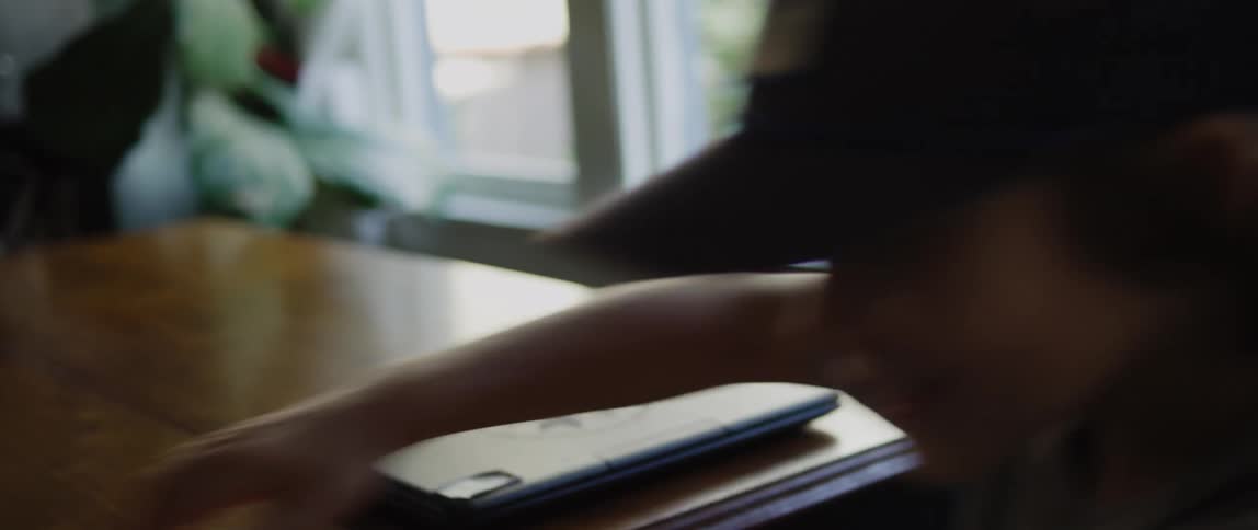 A boy wearing a baseball cap is sitting at a table looking at a laptop computer and he closes it and leaves