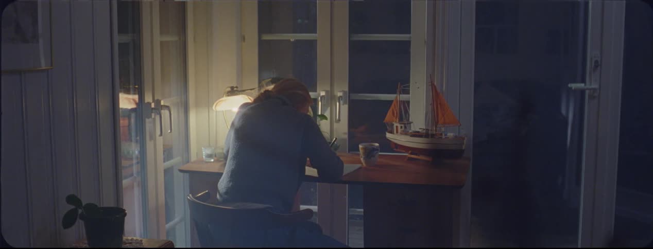 Woman walks over to a desk and sits in a chair and begins to write in a notebook