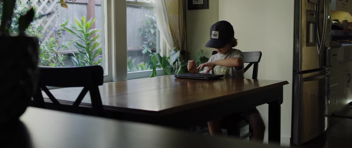 A boy wearing a baseball cap walks into a room and sits at a table in front of a laptop computer and he opens it and looks at the screen