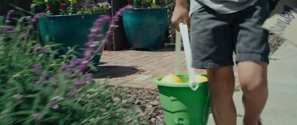 A child walks through a neighborhood carrying a bucket of lemons and a cardboard sign