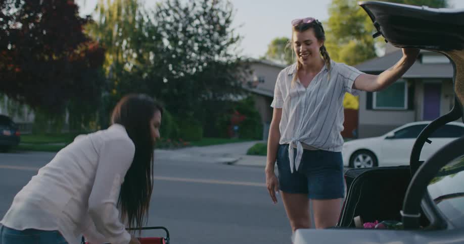 A woman takes her groceries and wagon out of the trunk of a car and waves goodbye to her friend