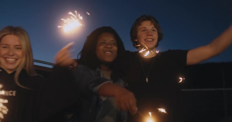 A group of friends wave sparklers together while standing outdoors in the late evening