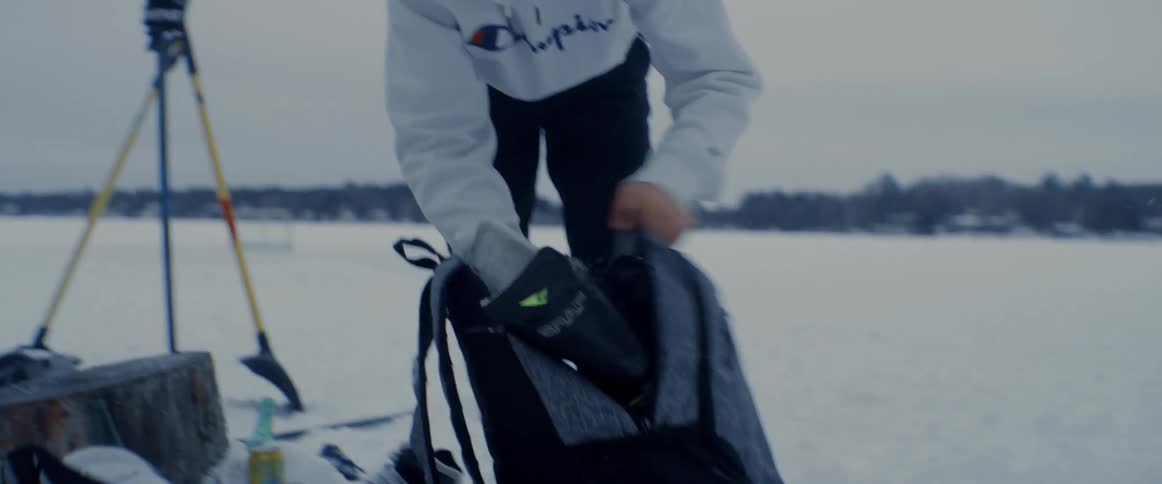 A teenage male hastily packs his ice skates into a backpack while outdoors