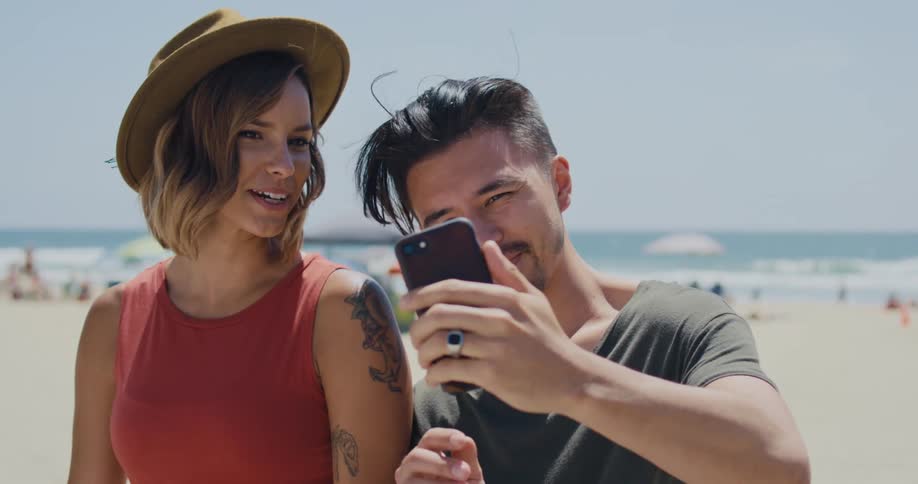 A man holding his phone up to capture a photo of him and his girlfriend at the beach