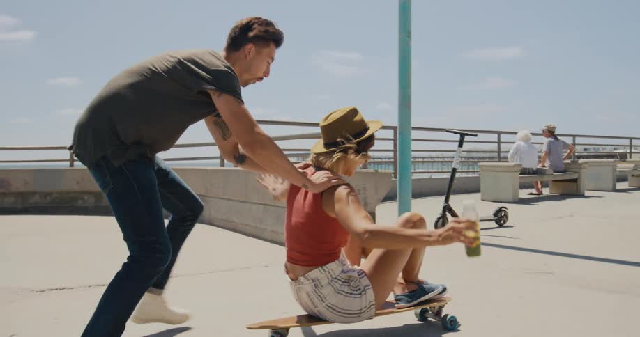 A girl is sitting on a skateboard holding a bottle and a man is pushing her across the concrete