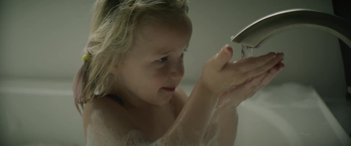 A young girl in a bath tub laughs as she puts her hands under the water coming out of the faucet