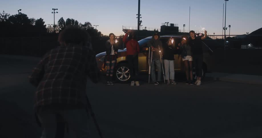 Photographer using tripod takes picture of friends posing against car with silhouette of stadium in the background