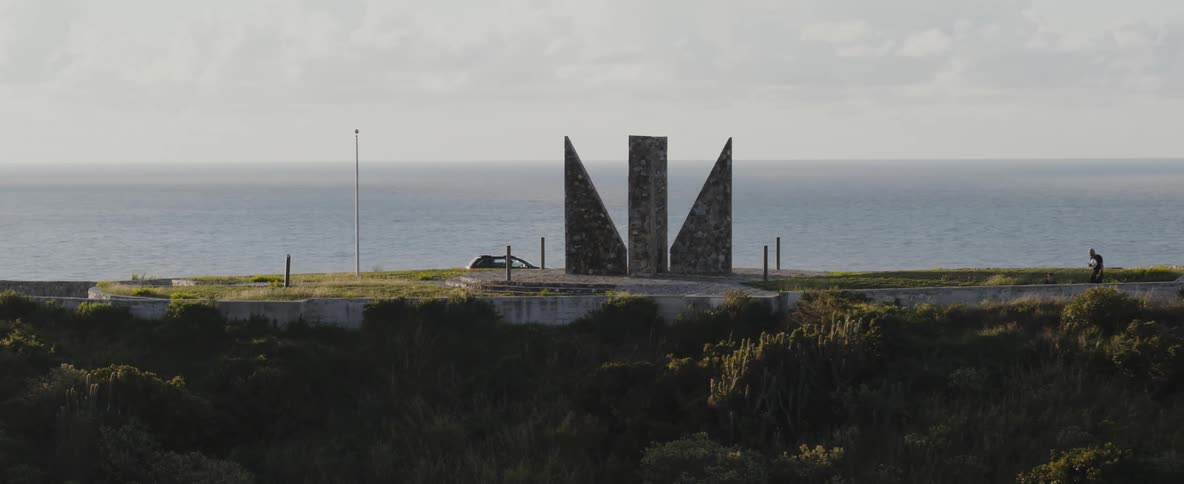 A person walks along the shore towards a monument as a car drives by