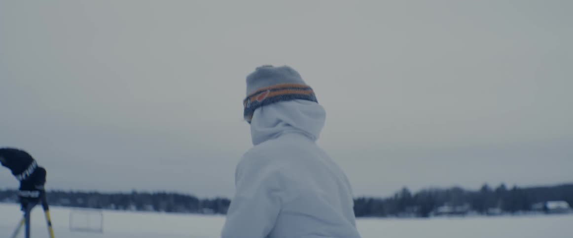 Woman wearing a jacket and a toque zips up a backpack then picks up a hockey stick and begins to leave the snowy field