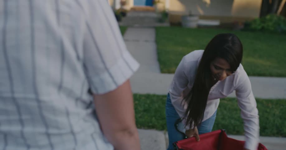A delivery person opens the trunk of a car and a woman takes out a cart and puts a bag of groceries in and waves good bye and walks towards her home