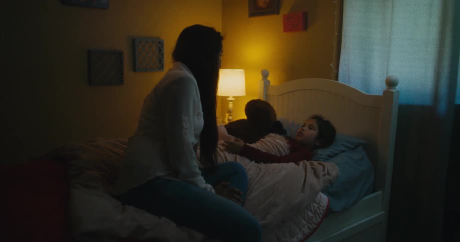 Mother with long straight hair blue shirt and jeans sits on bed talking to daughter lying in bed in bedroom at night