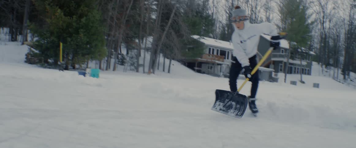 A child shovels snow while ice skating in a small town on a winter day