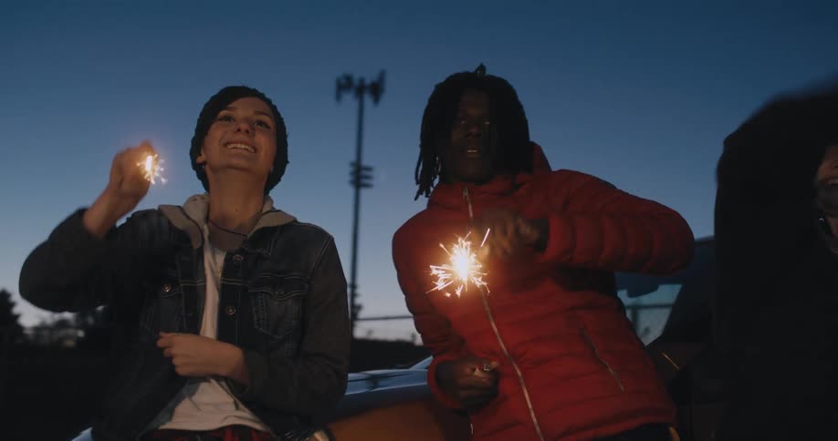 Friends stand against a car waving sparklers at night