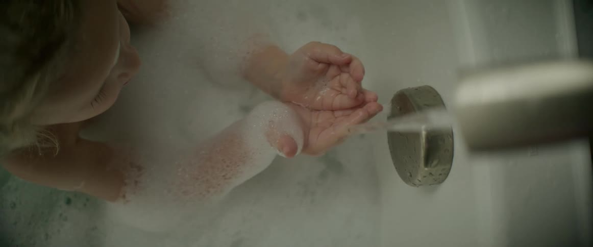 A young girl sitting in a soapy bathtub uses water from the faucet to wash the soap from her hands