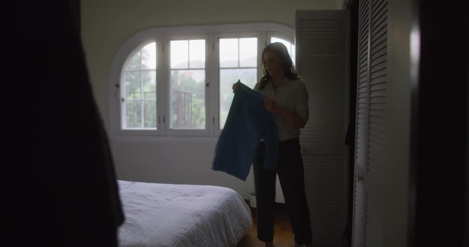 A woman walks into a bedroom and takes a shirt out of the closet and puts it on the bed