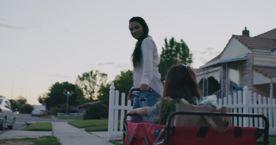 A mom looks back at her daughter smiling and making faces as she pulls her and groceries down the sidewalk