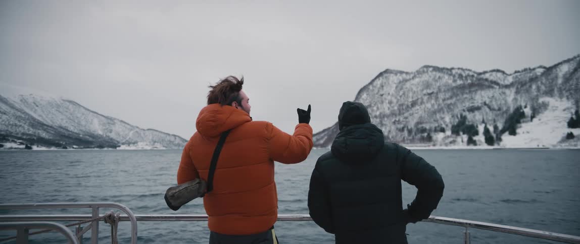 Two people stand on a boat and point at the snowy mountains ahead of them