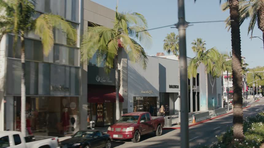 Cars travel on the tree lined street in front of the retail buildings as pedestrians walk down the sidewalk